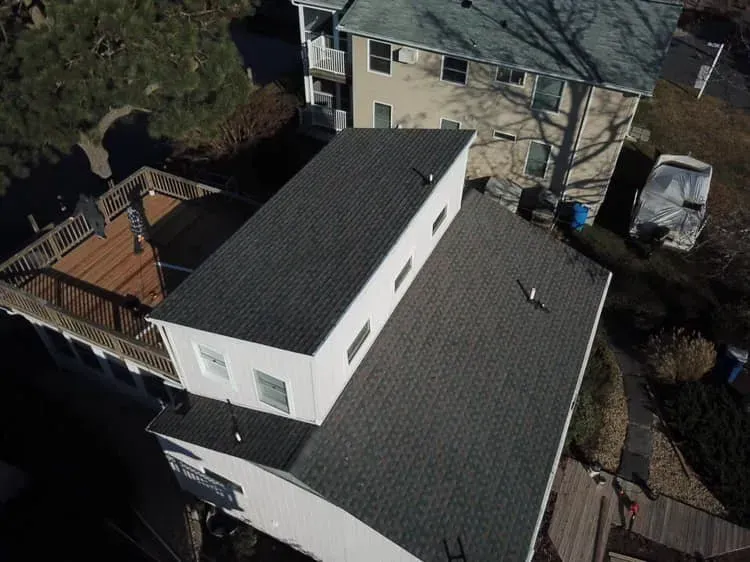 Aerial view of a white house with dark roof, deck, and surrounding trees and driveway