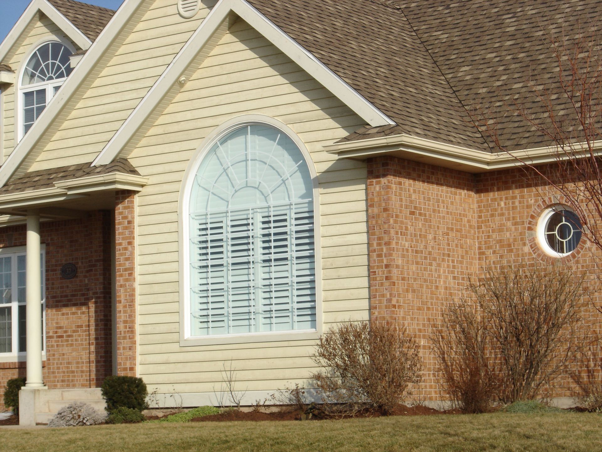 Front exterior of a house with beige siding, brick walls, arched window, and shrubs along the foundation
