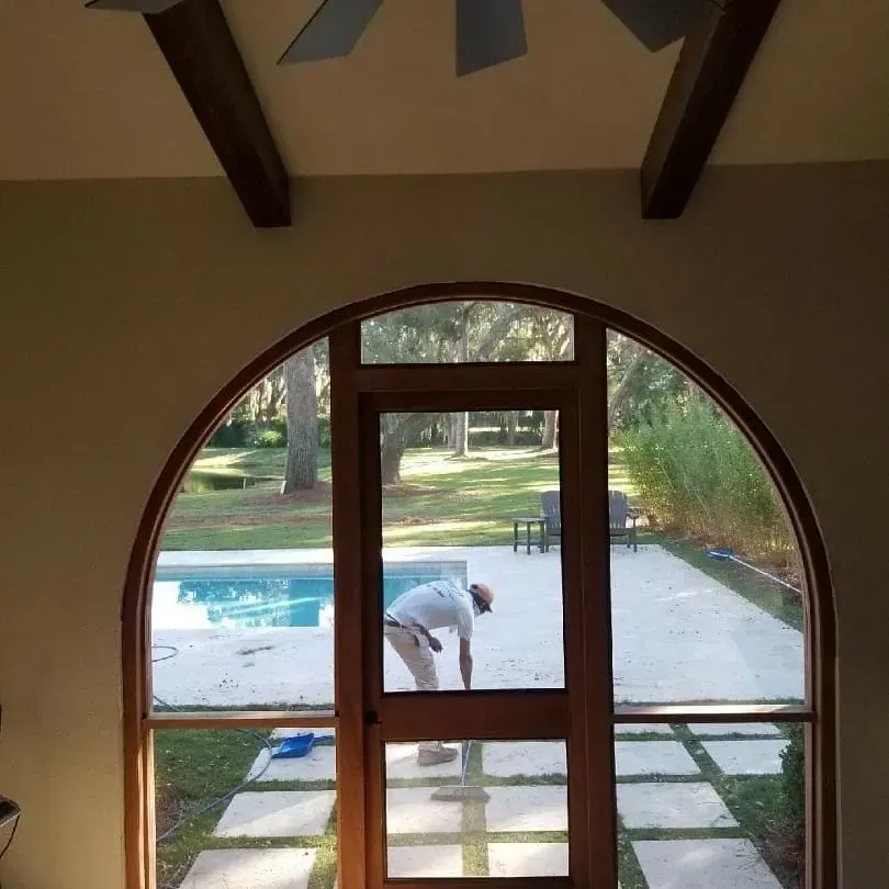 Indoor view through arched window of a backyard pool where a person is bending near the patio.