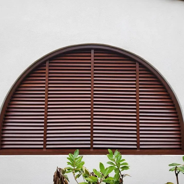Semi-circular brown window shutters on a white wall with green plants below