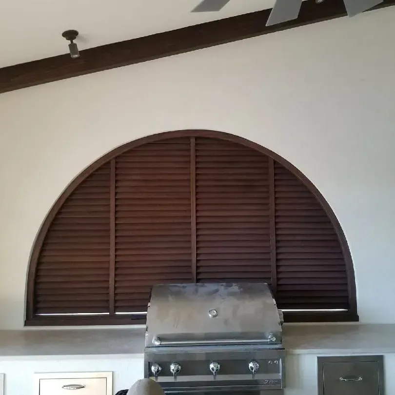 Arched window with closed brown shutters above a stainless steel grill on a white wall