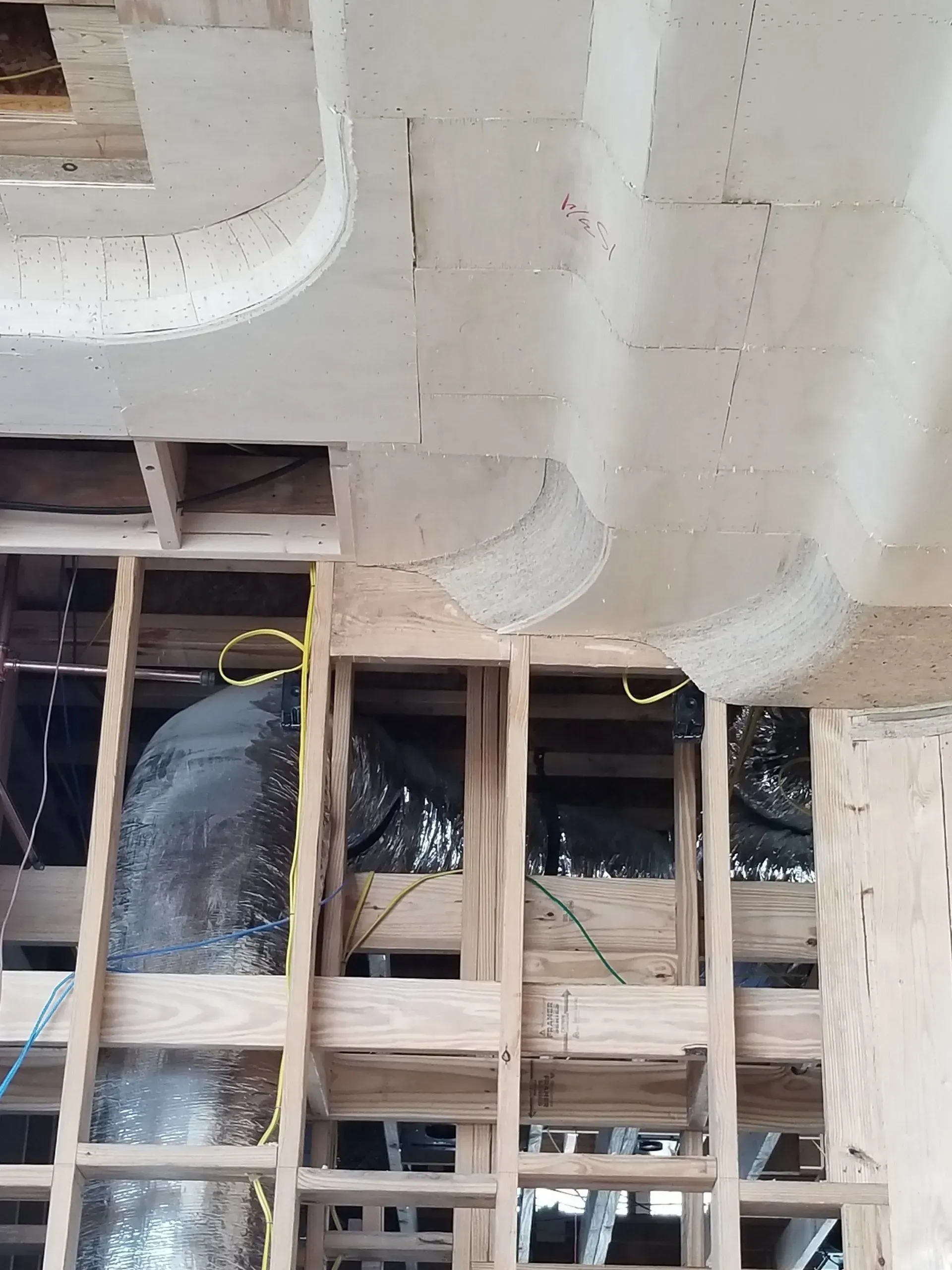 Exposed wooden framing and ducts inside a room under construction with white ceiling panels and black insulation.