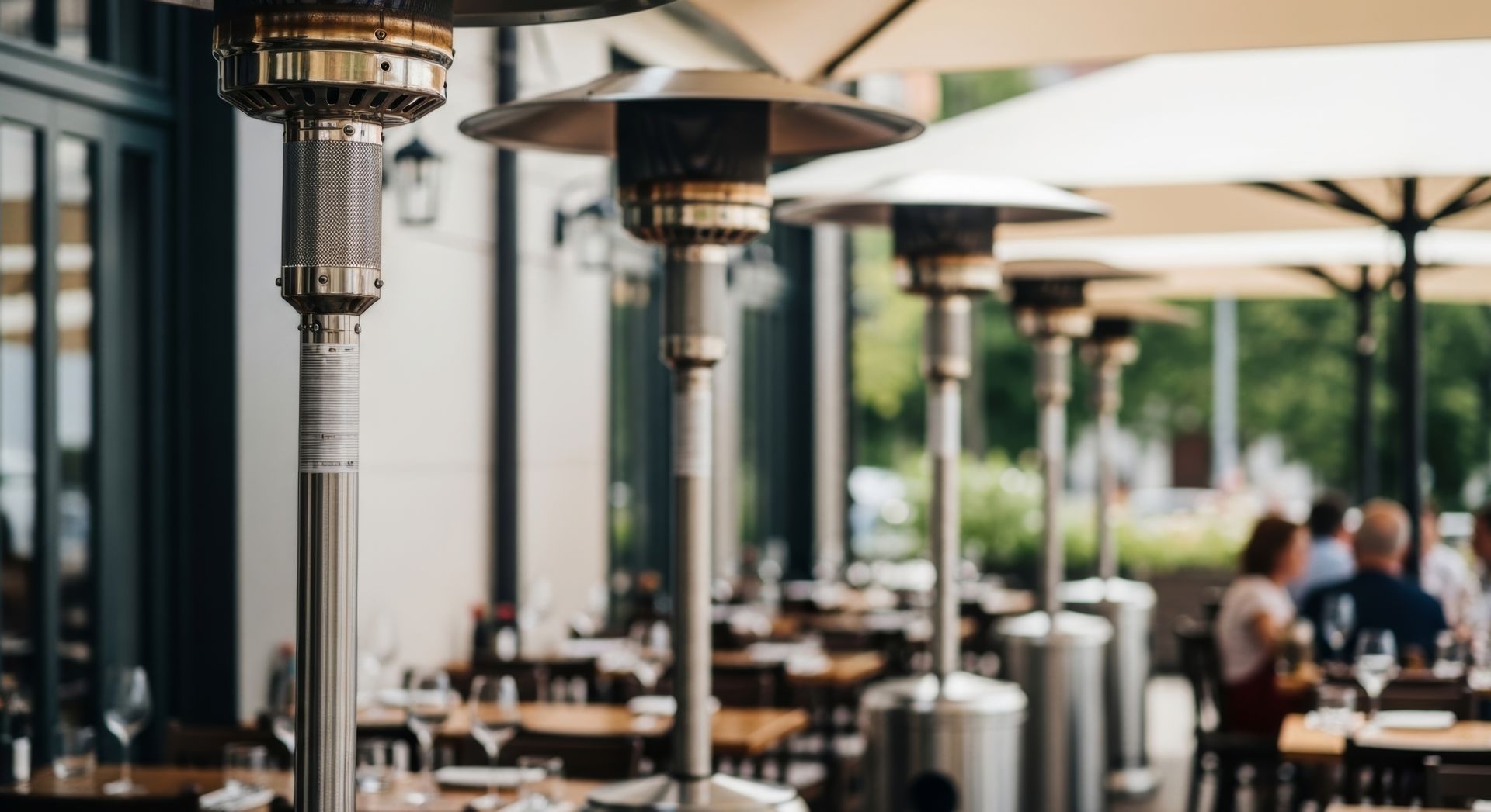 Outdoor café patio with metal tables, large umbrellas, and heat lamps, with people seated in the background