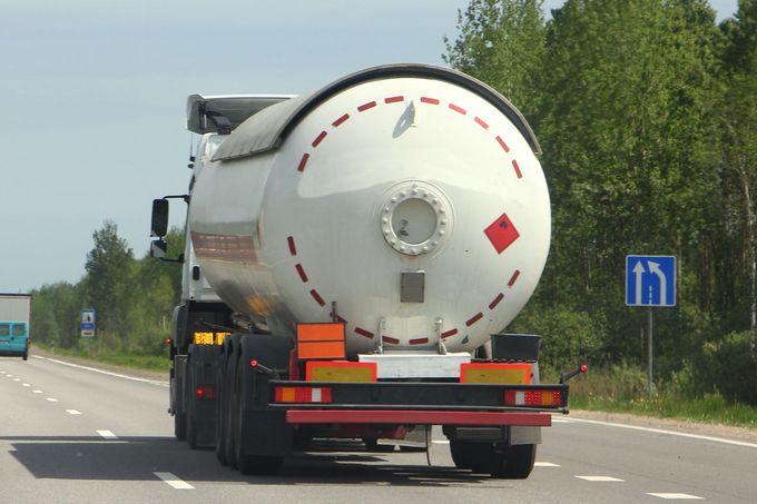 White tanker truck driving on a highway, viewed from behind, with trees and road signs along the road