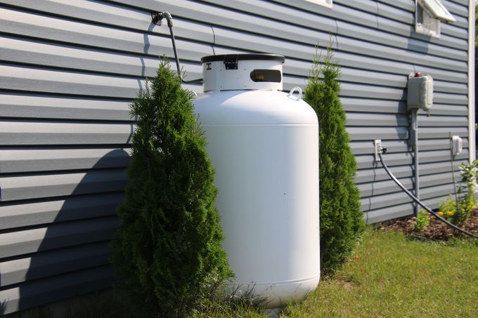 White propane tank beside a gray house wall, partly hidden by green shrubs.