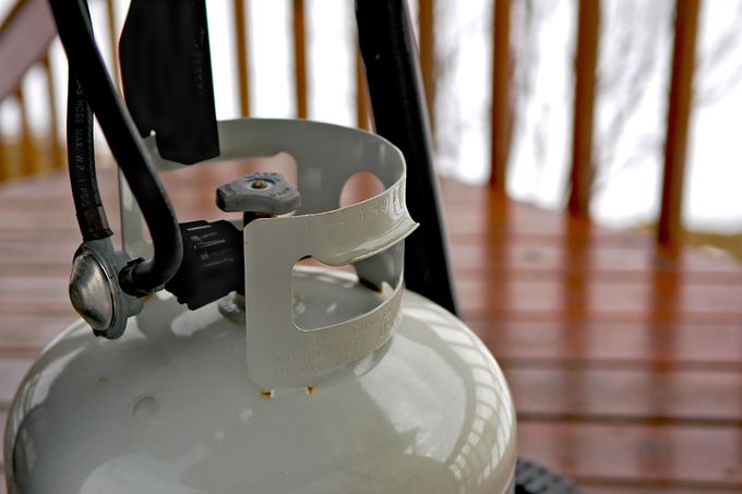 Close-up of a white propane tank with a black valve and hose indoors on a wooden floor