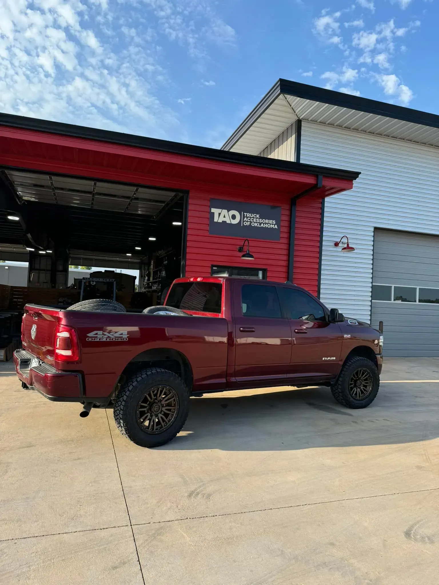 Red lifted pickup truck parked outside a red and white auto shop under a blue sky.