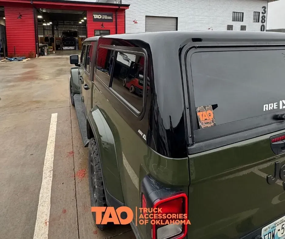 Olive-green Jeep parked outside a truck accessories shop with a black roof cover.