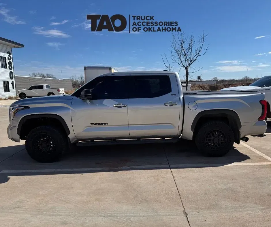Silver pickup truck parked on a lot under a clear sky, with a TAO Trucks Accessories Oklahoma sign in the background