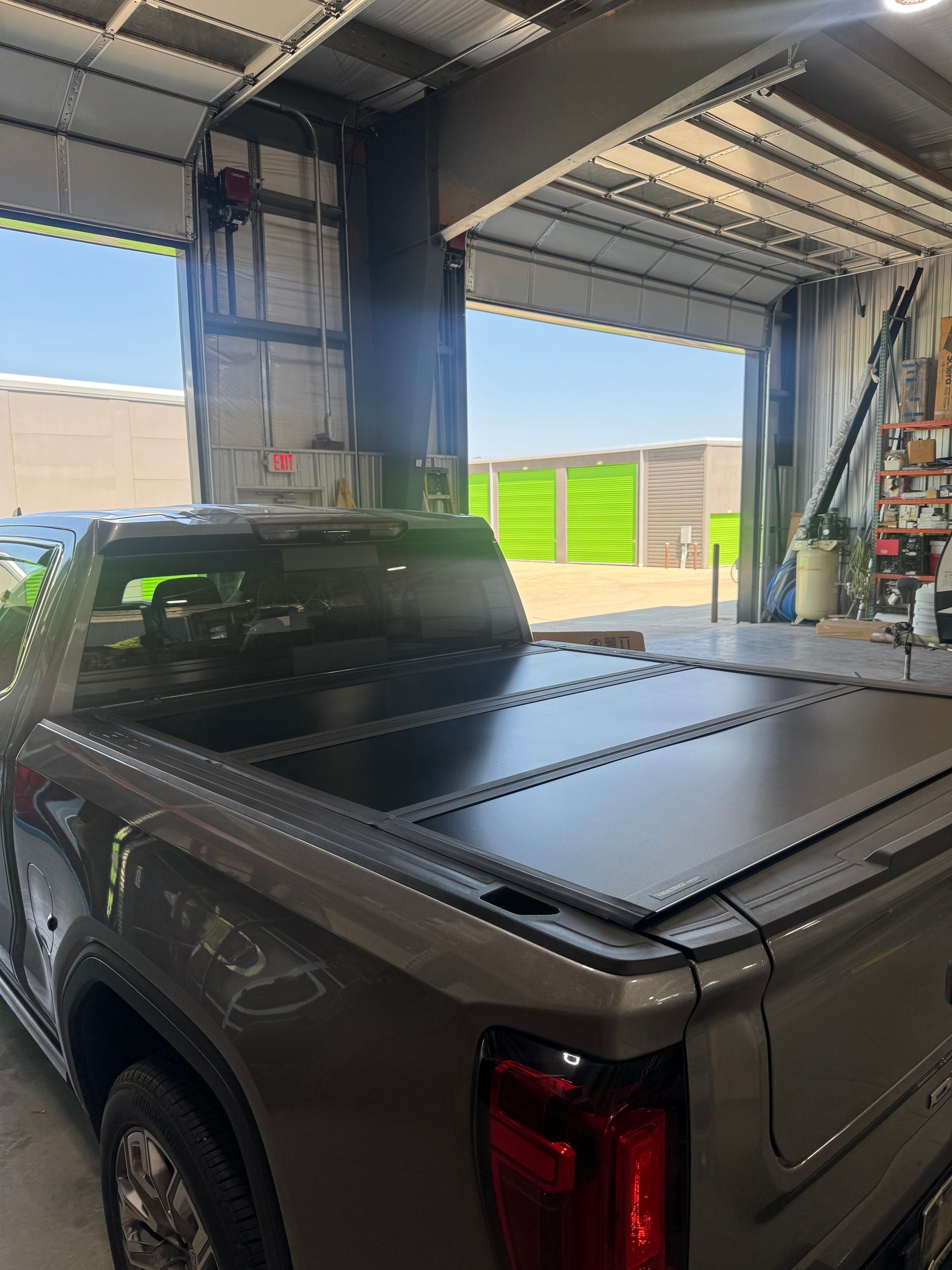 Gray pickup truck parked inside an industrial garage with a covered bed and open bay doors.