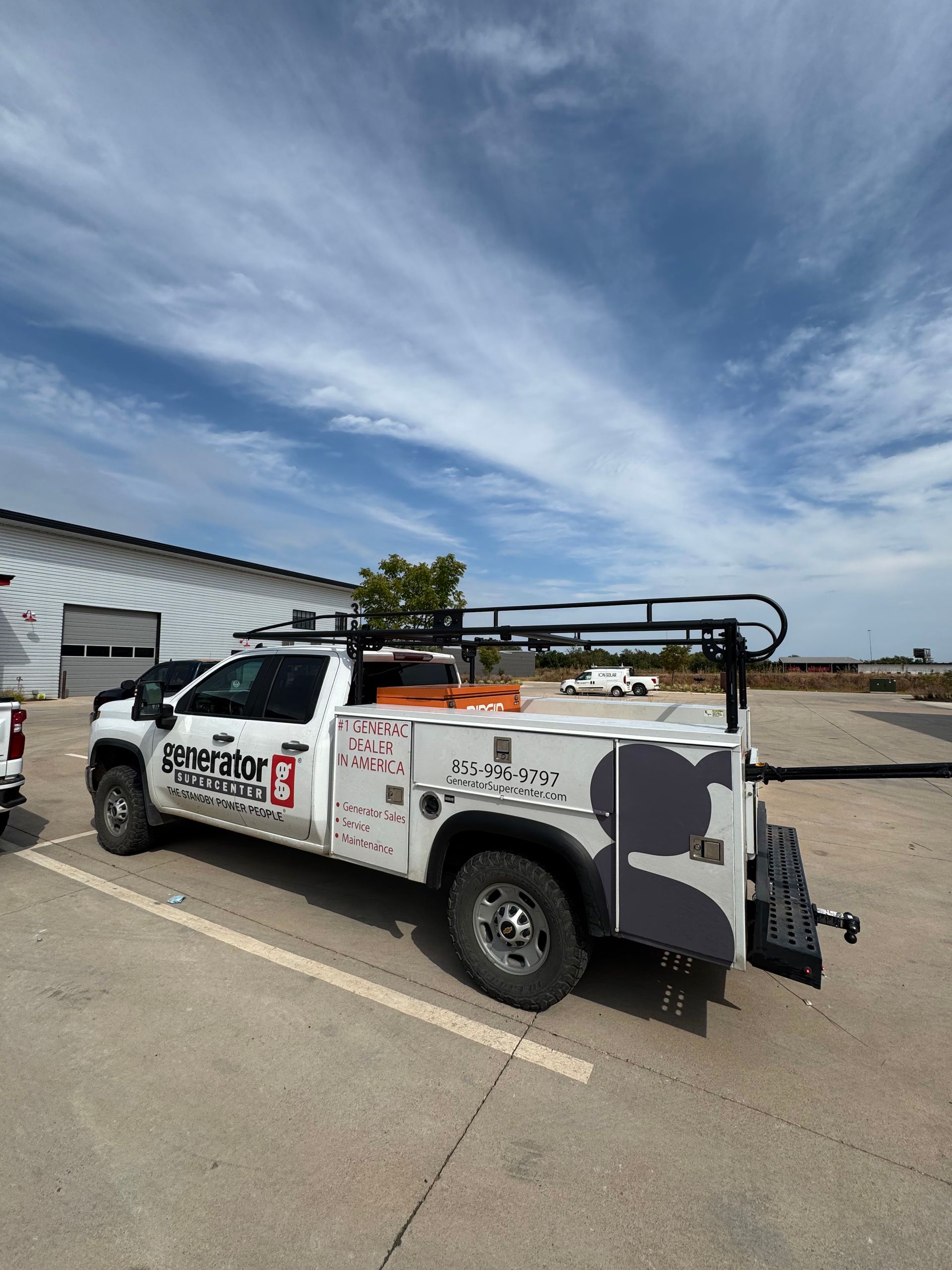 White utility truck with roof rack parked on a lot beside a warehouse under a blue sky.