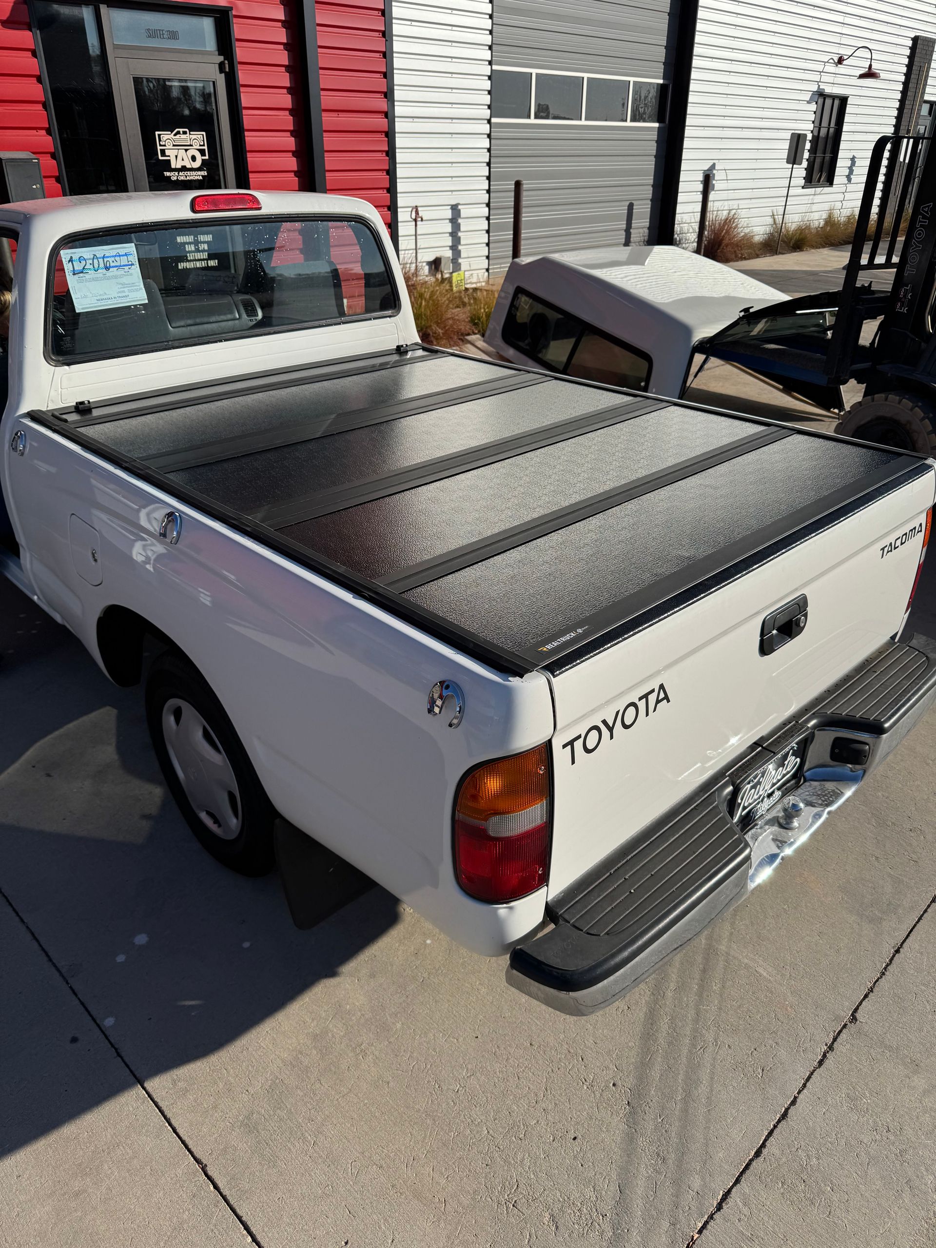White Toyota pickup truck parked outside a building on a sidewalk, with a black bed cover.