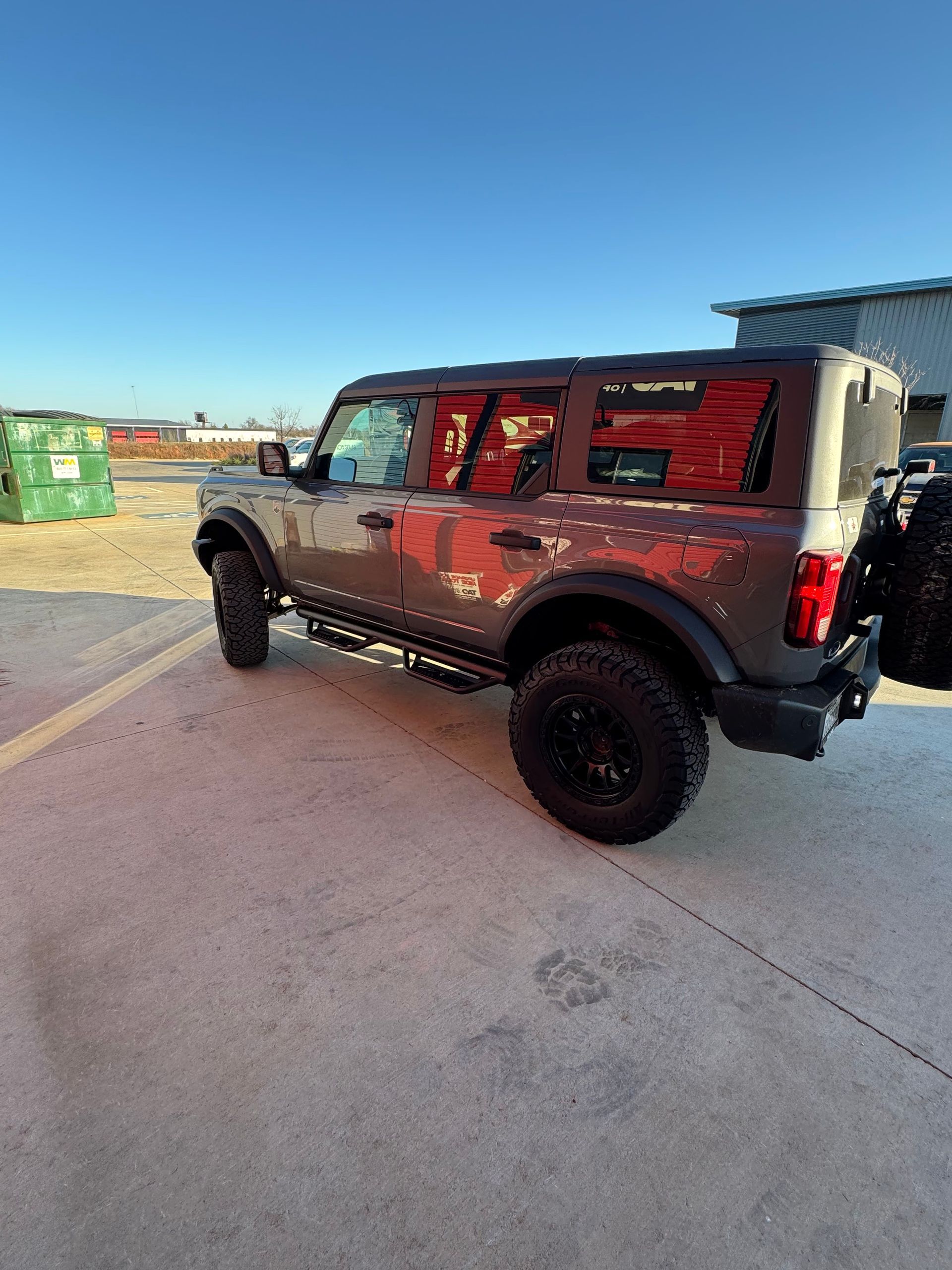 Silver Jeep Wrangler with red reflections parked on a concrete lot under a blue sky.
