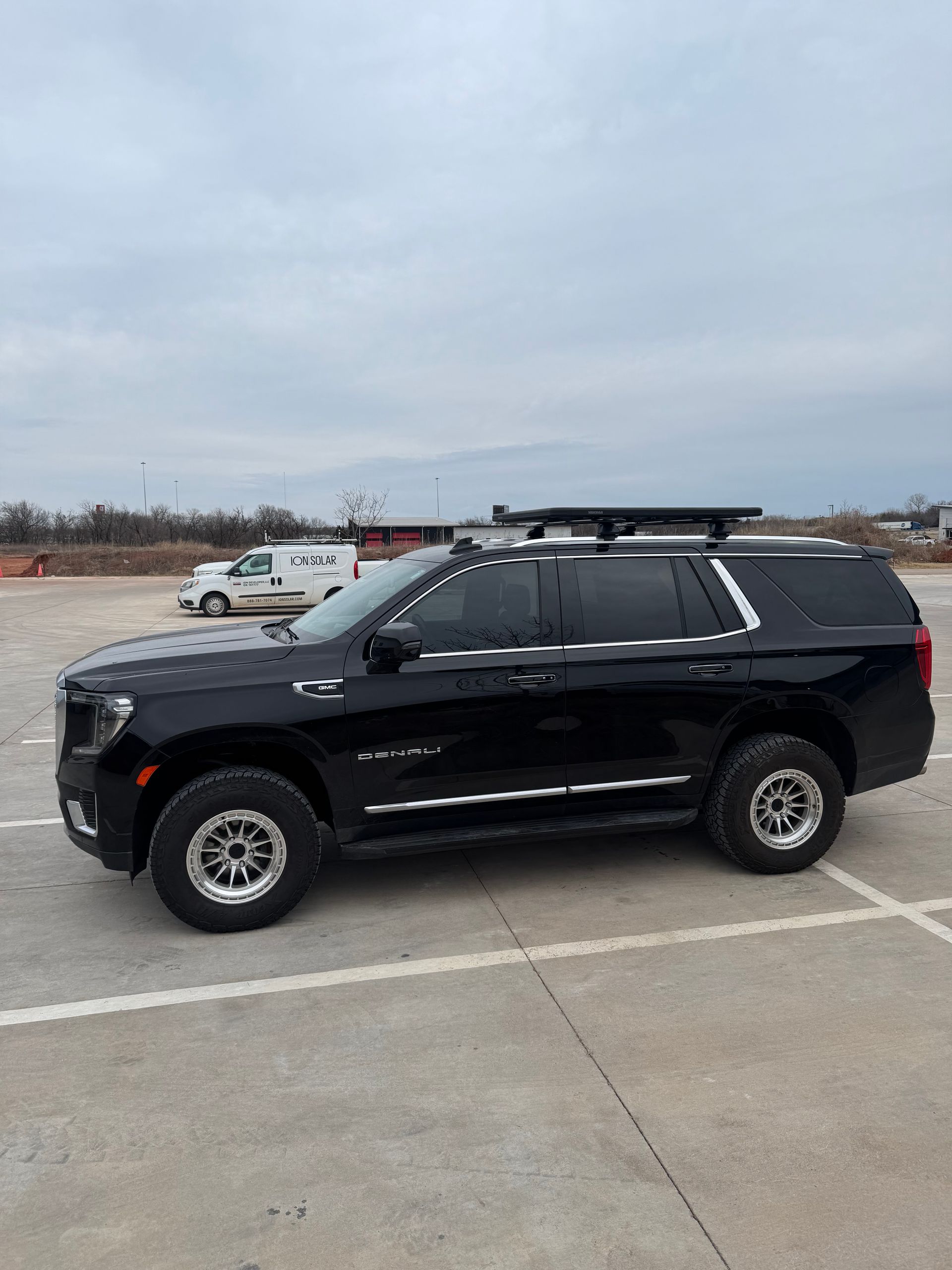 Black SUV parked on a concrete lot under a cloudy sky, side view with roof rack