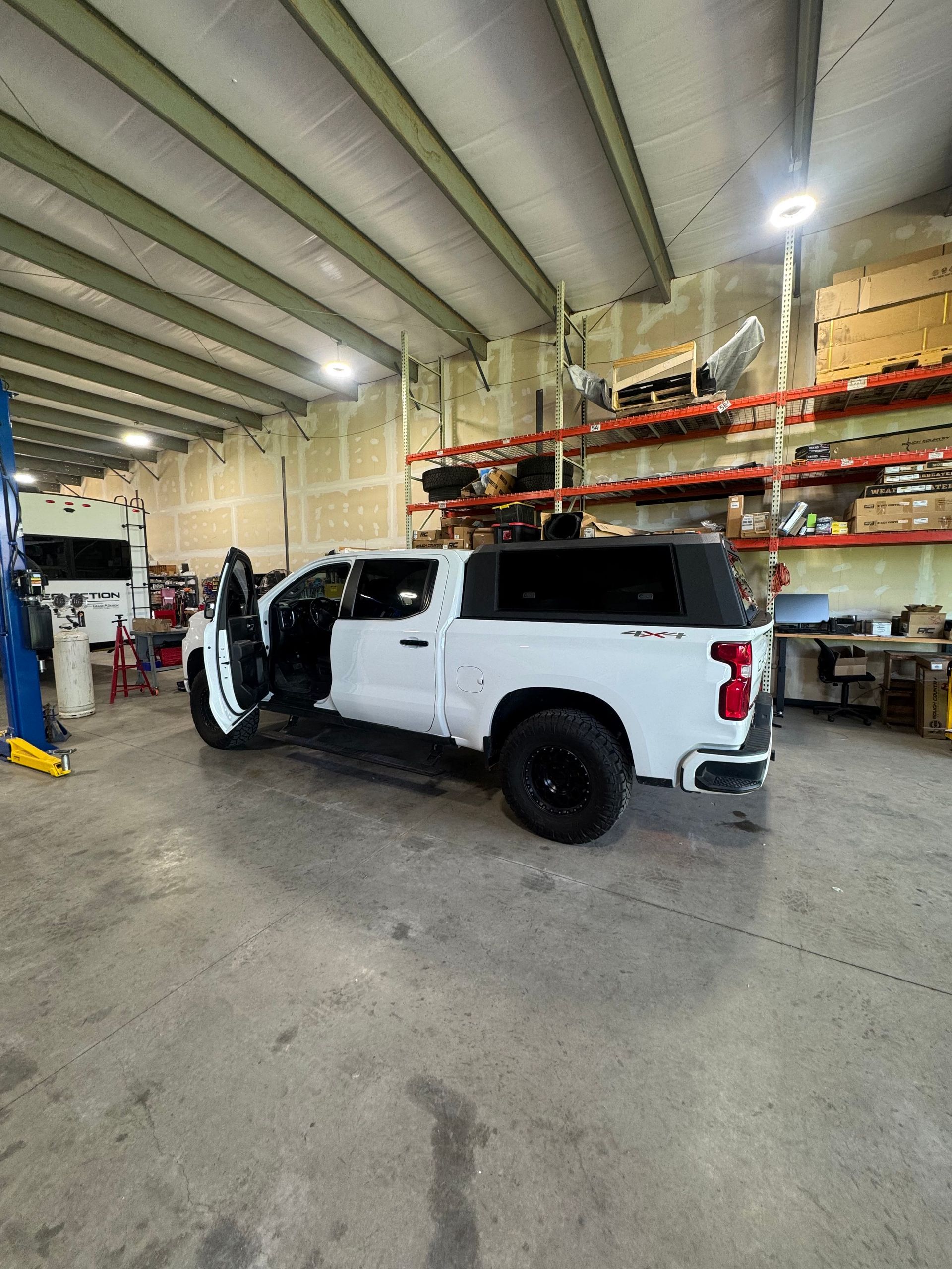 White pickup truck in an auto shop with its front door open, under high warehouse shelving.