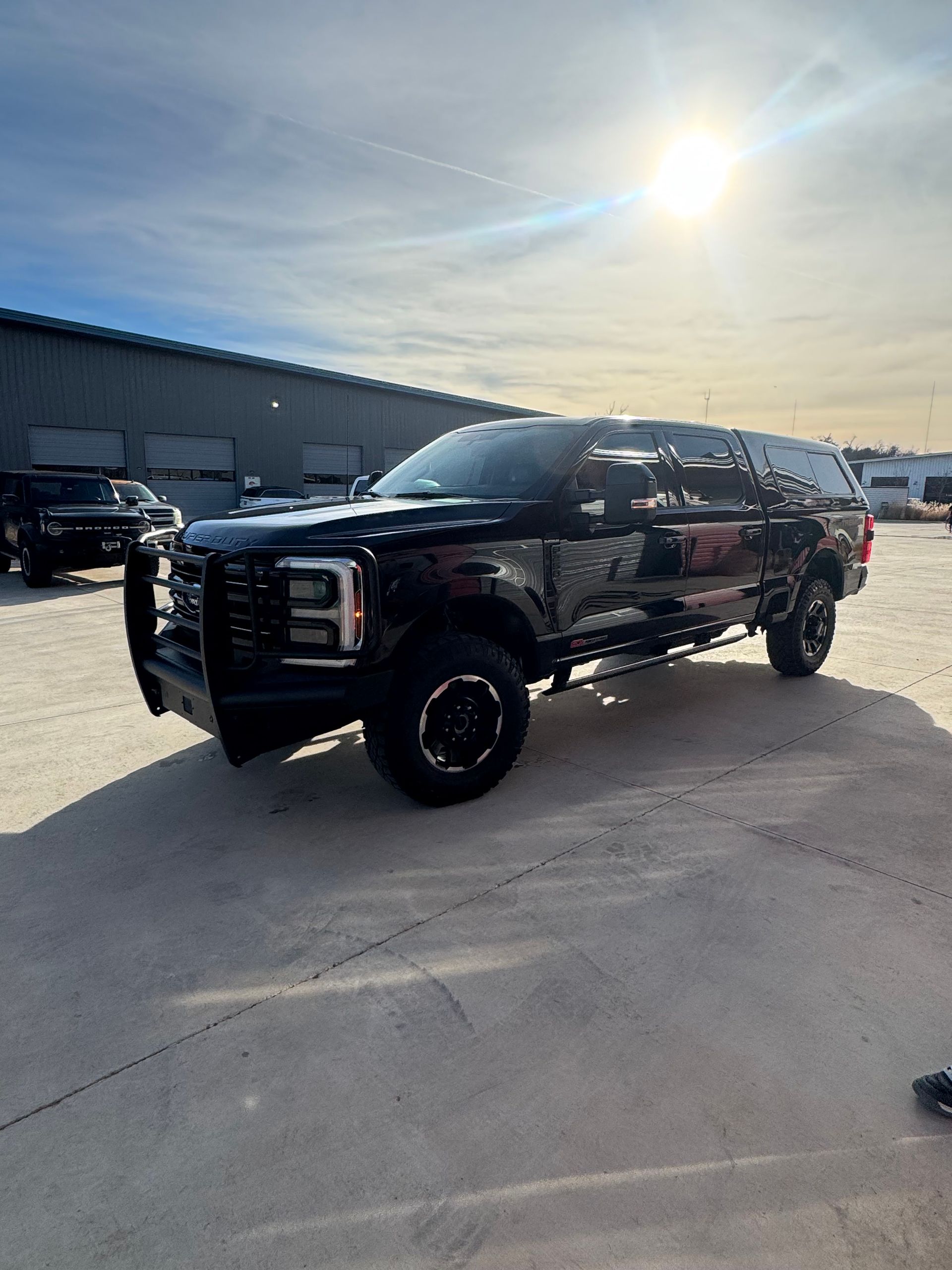 Black lifted SUV in a snowy lot under bright sun, with a warehouse in the background.