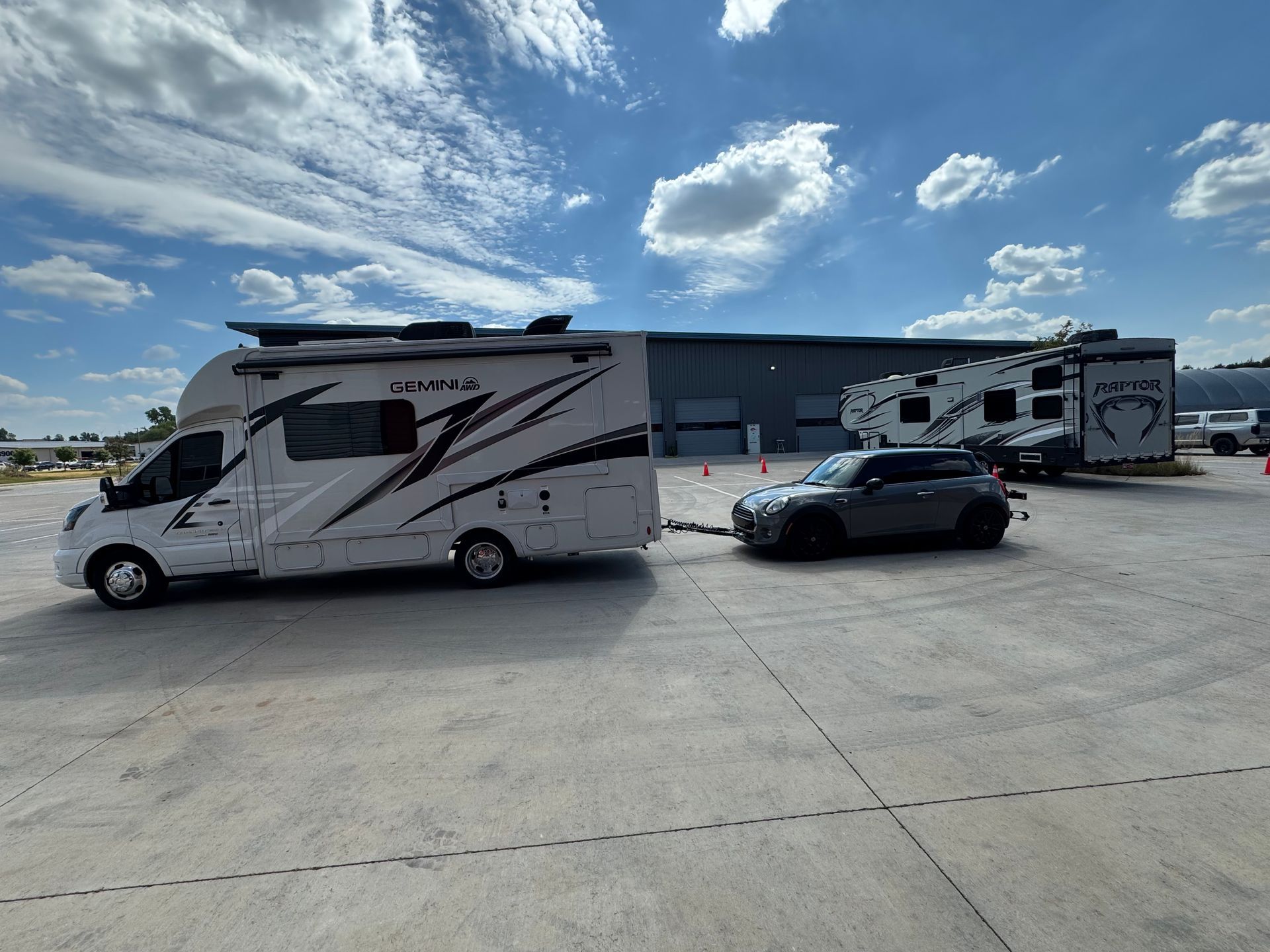 Motorhome, sports car, and RV trailer parked on a wide concrete lot under a partly cloudy sky.