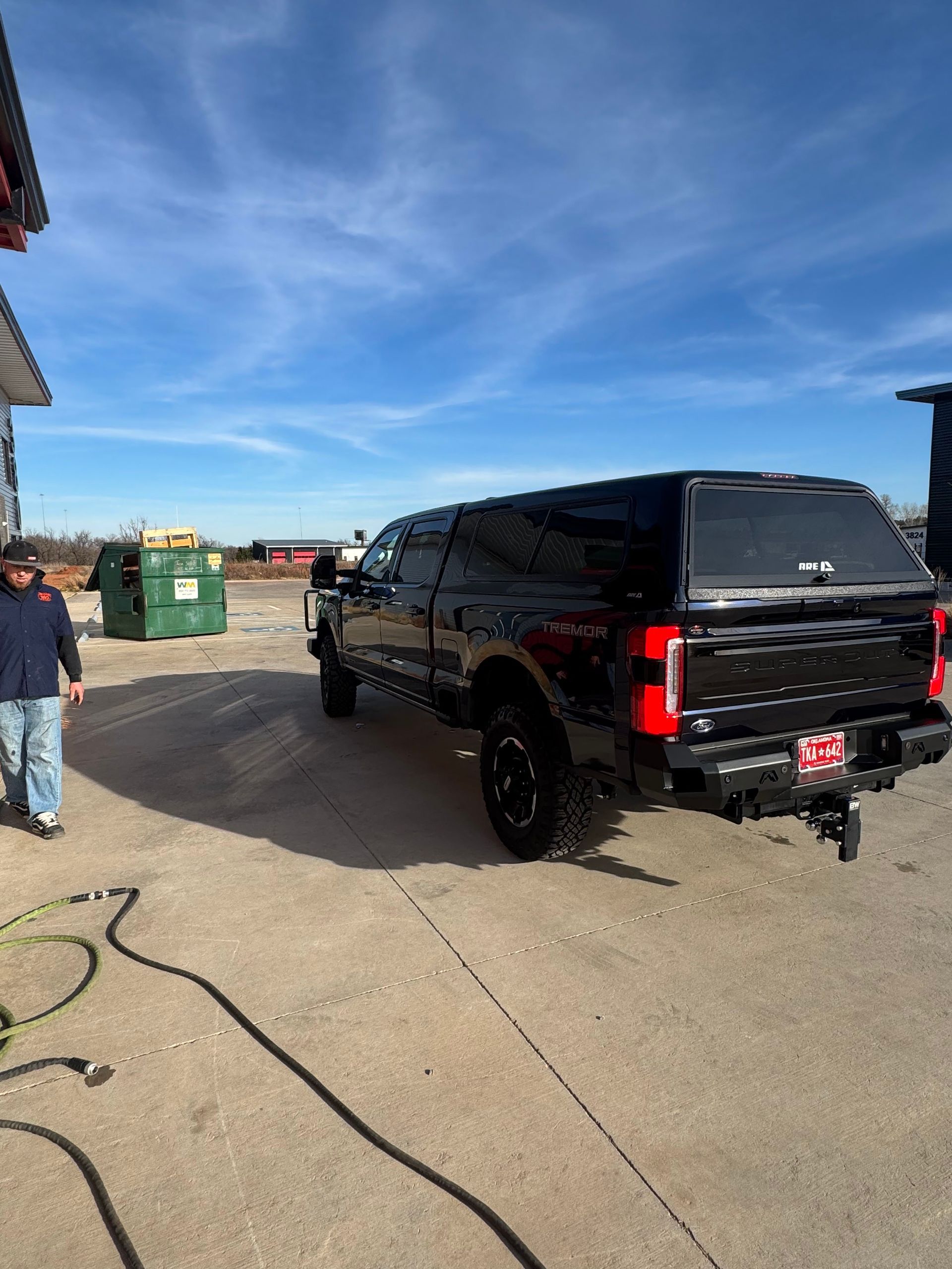 Black pickup truck parked on a lot beside a hose, with a person standing near green dumpsters under a blue sky.