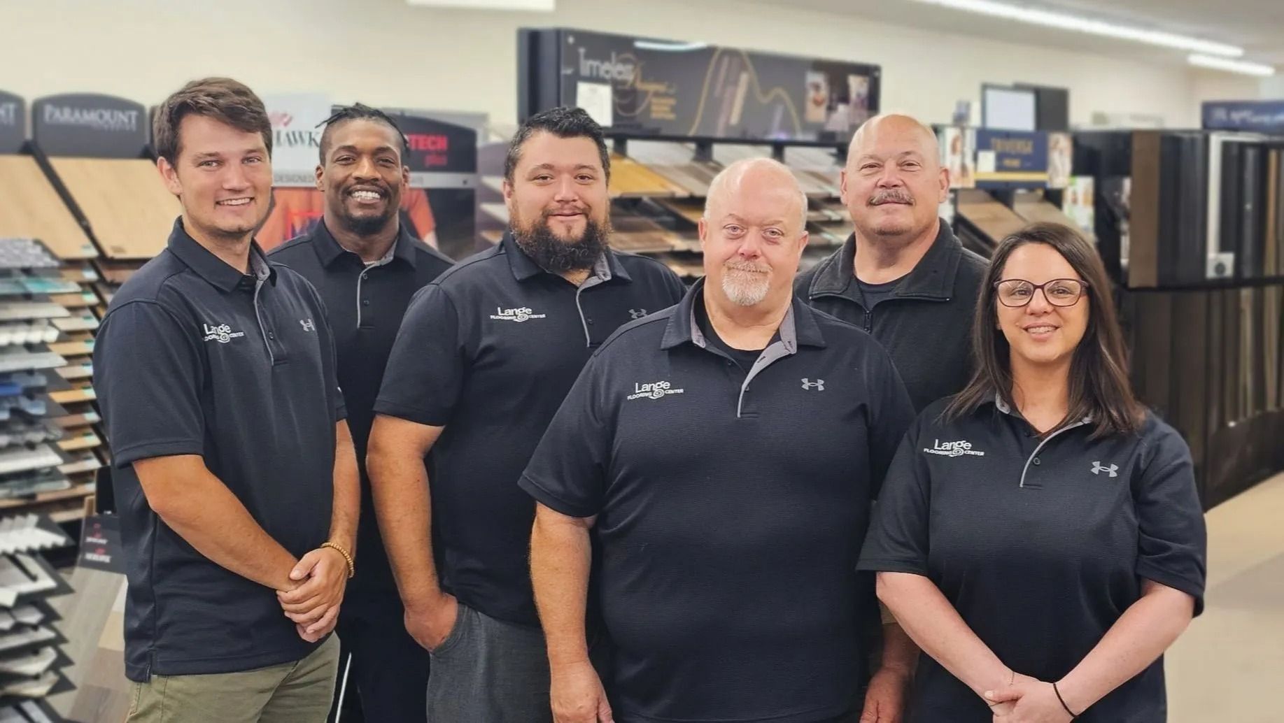 Six people in black polo shirts standing inside a store; the backdrop shows flooring samples.