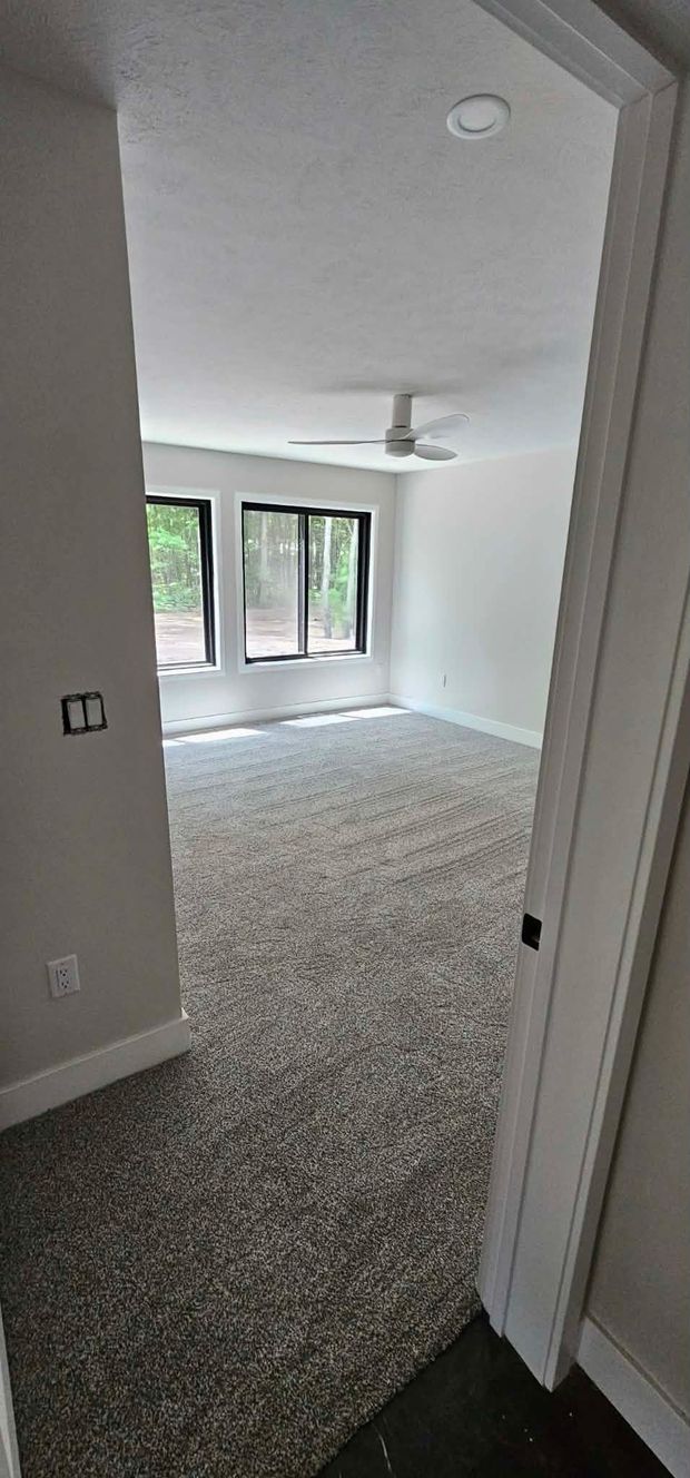 View into a carpeted bedroom with white walls, a ceiling fan, and windows with black frames.
