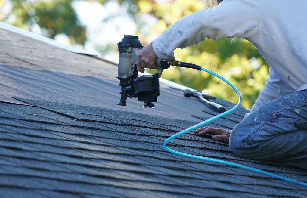 Worker using a nail gun to install shingles on a roof with a blue air hose