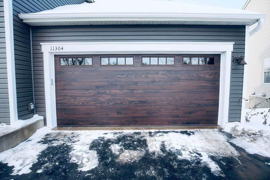 A garage door with a wooden door and a snowy driveway in front of a house.