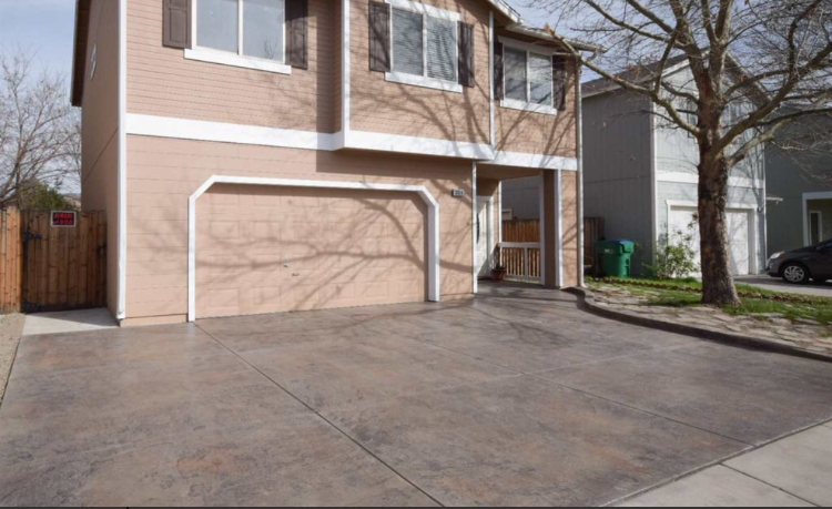 Tan two-story house with a brown concrete driveway, garage, and a tree casting shadows.