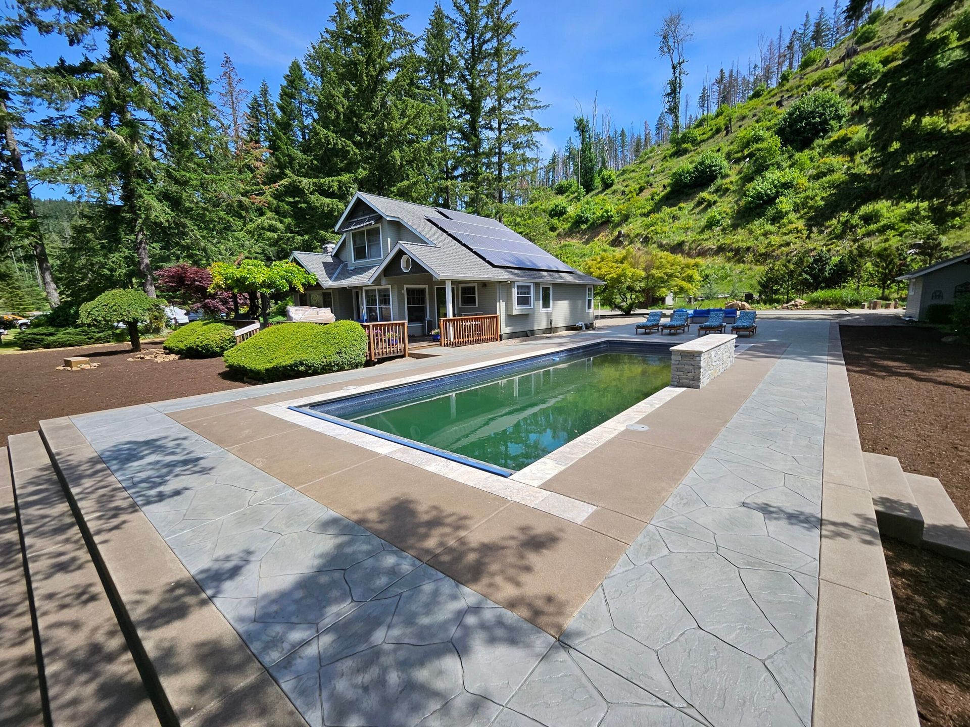 A house with a pool in a wooded area; blue pool surrounded by a stone patio.