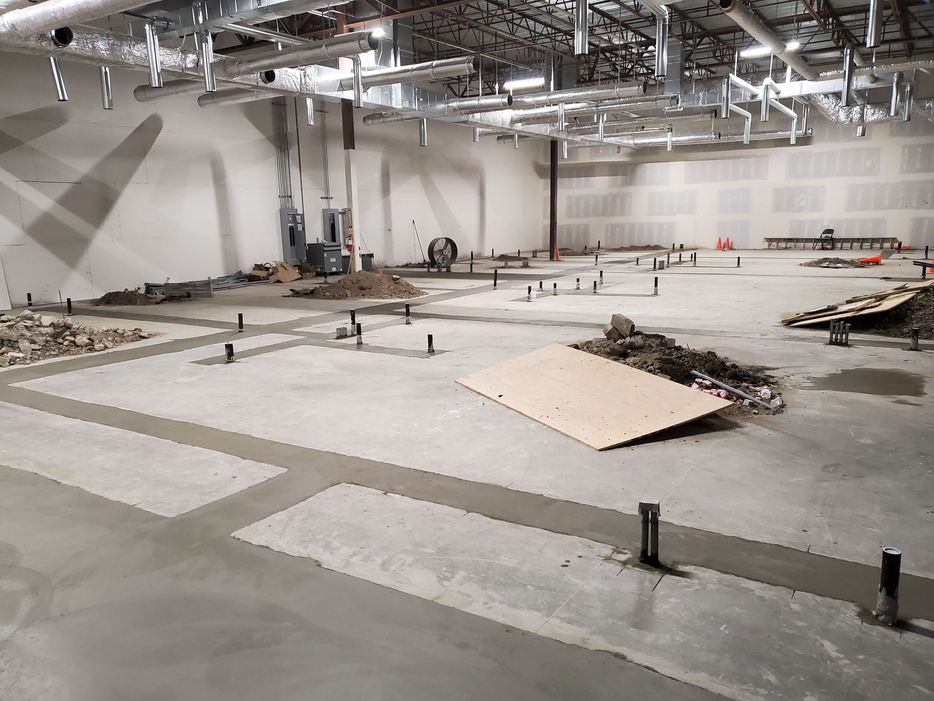 Interior construction site, concrete floor with pipes, ductwork, and debris.