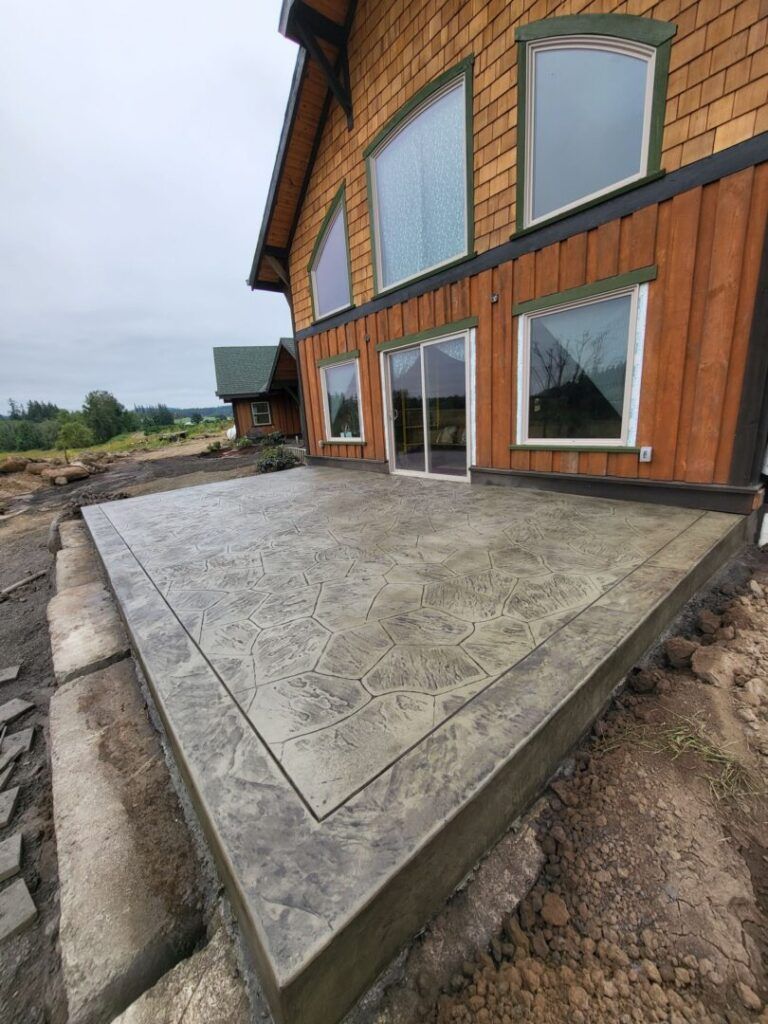 Newly poured and stamped concrete patio next to a house with wood siding and large windows.