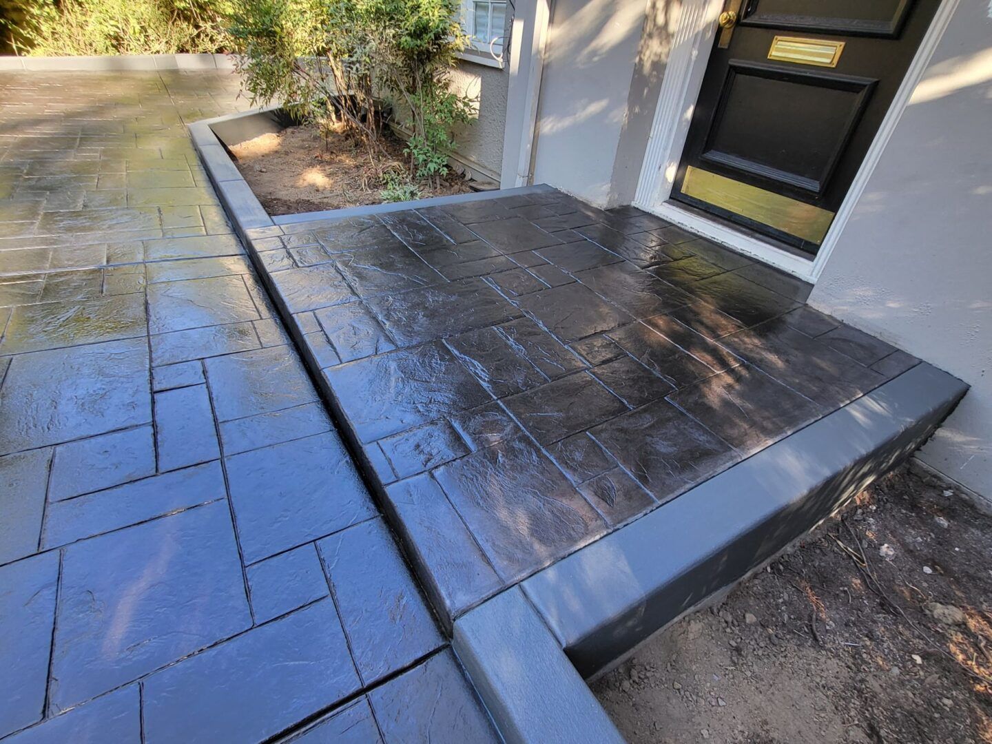 Dark gray stamped concrete patio with decorative stone pattern, leading to a front door with a small garden area.