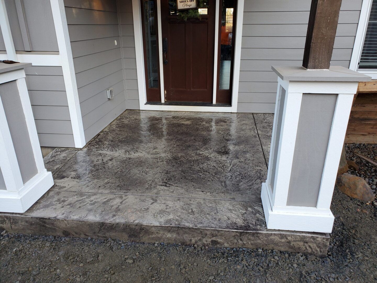Concrete front porch with two white and gray columns, a brown door, and gray siding.