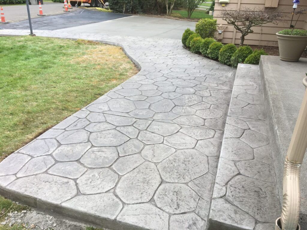 Stone-patterned concrete walkway and steps leading to a house. Green grass on the left, shrubs on the right.