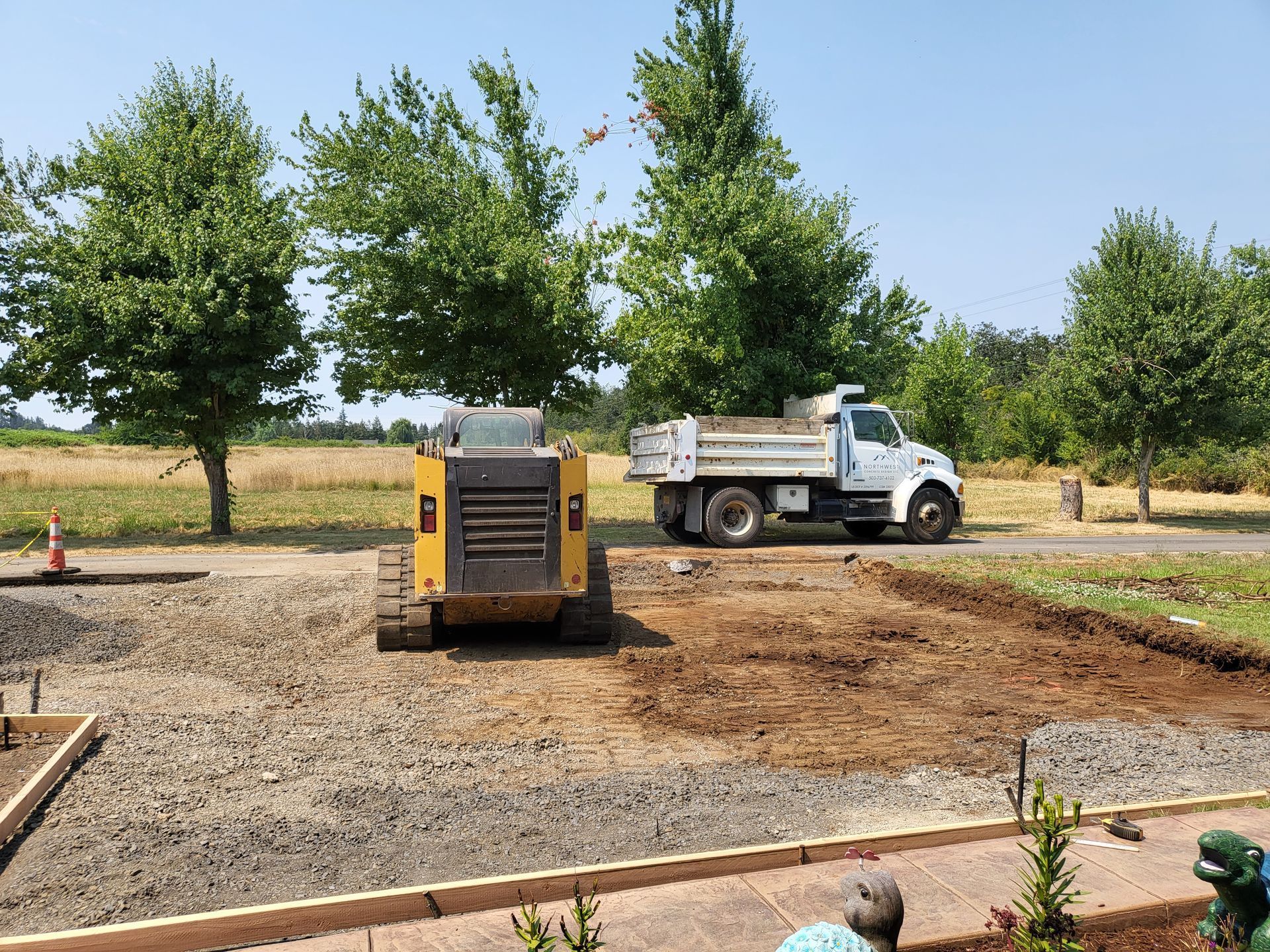 Skid steer and dump truck on gravel, preparing a ground area with trees and blue sky.