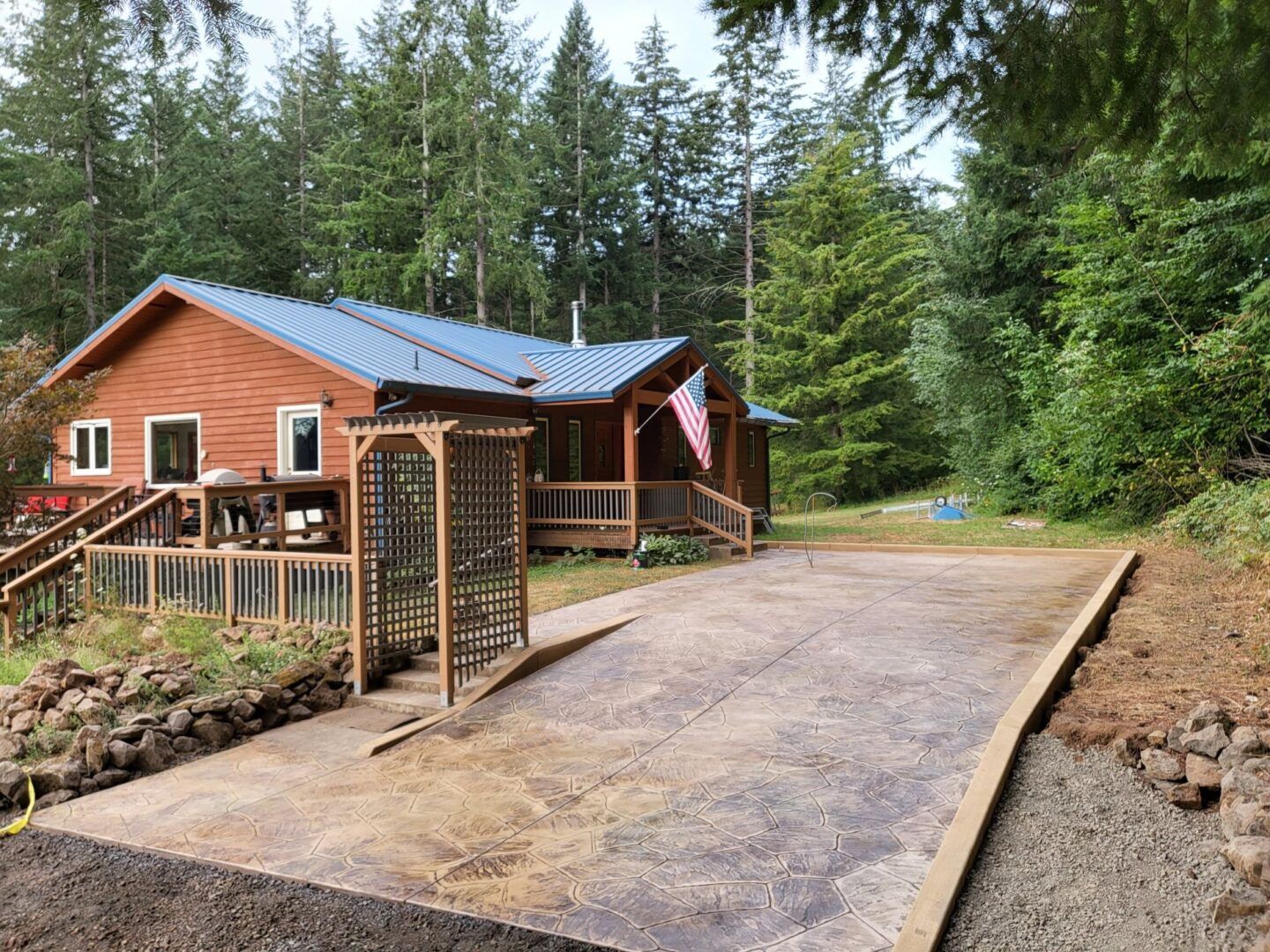 Brown house with blue roof, wooden deck, gravel driveway, and American flag. Forest backdrop.