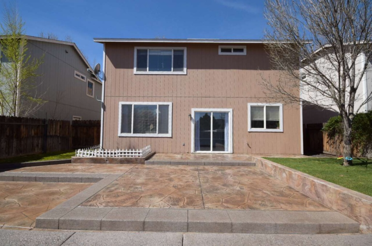 Back of a two-story beige house with a concrete patio. Sliding glass door and windows visible. Sunny day.