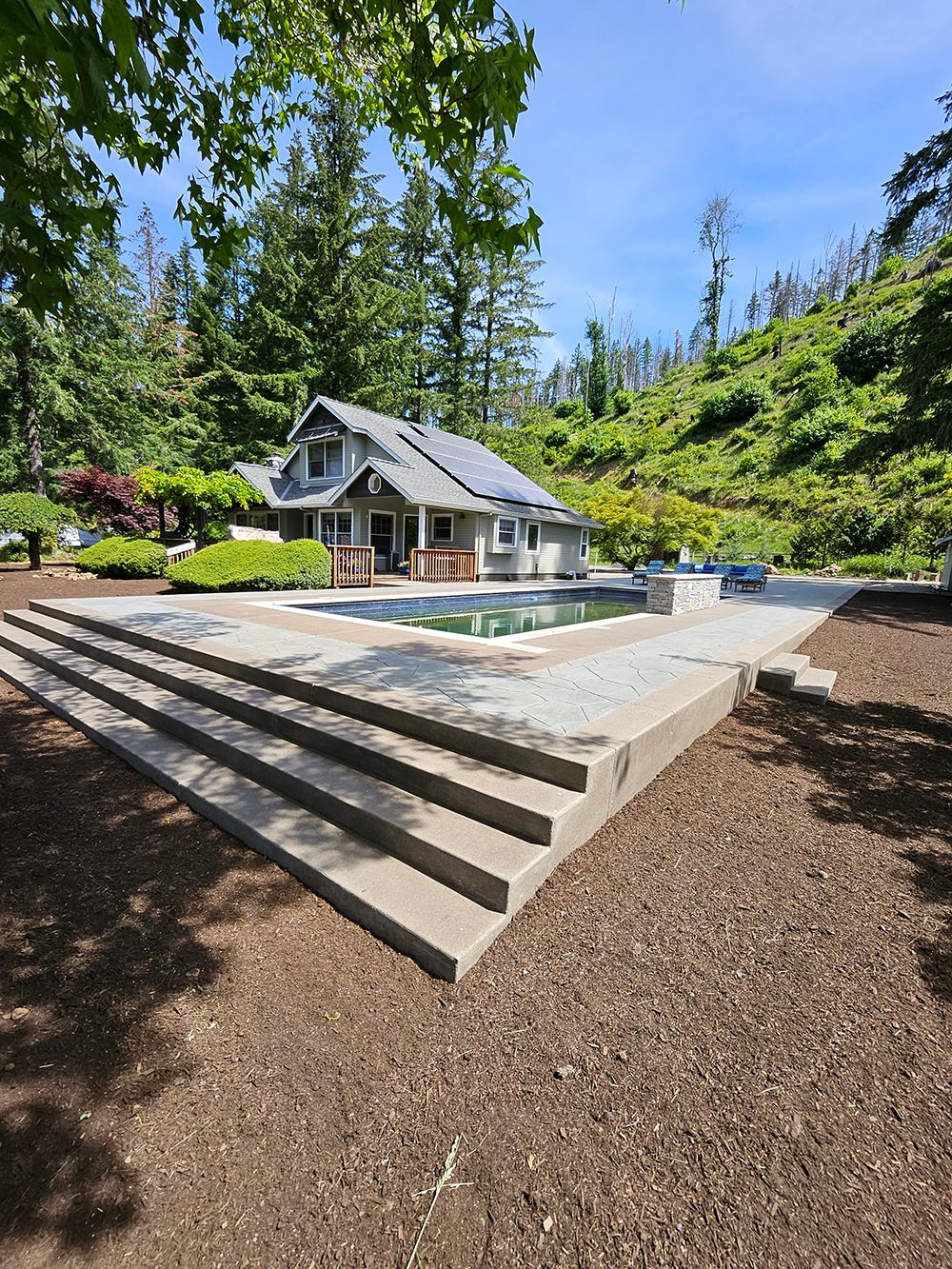 House with pool, steps, and wooded hillside on a sunny day.