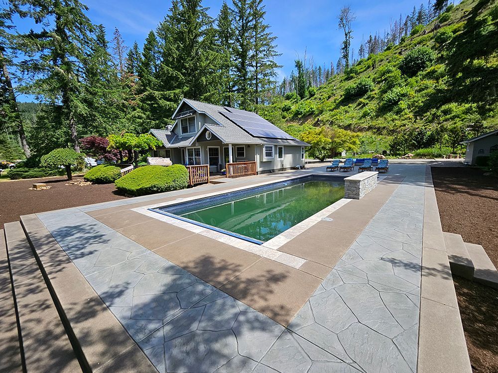 House with pool in a wooded setting; blue pool water, gray stone patio, green trees, sunny day.