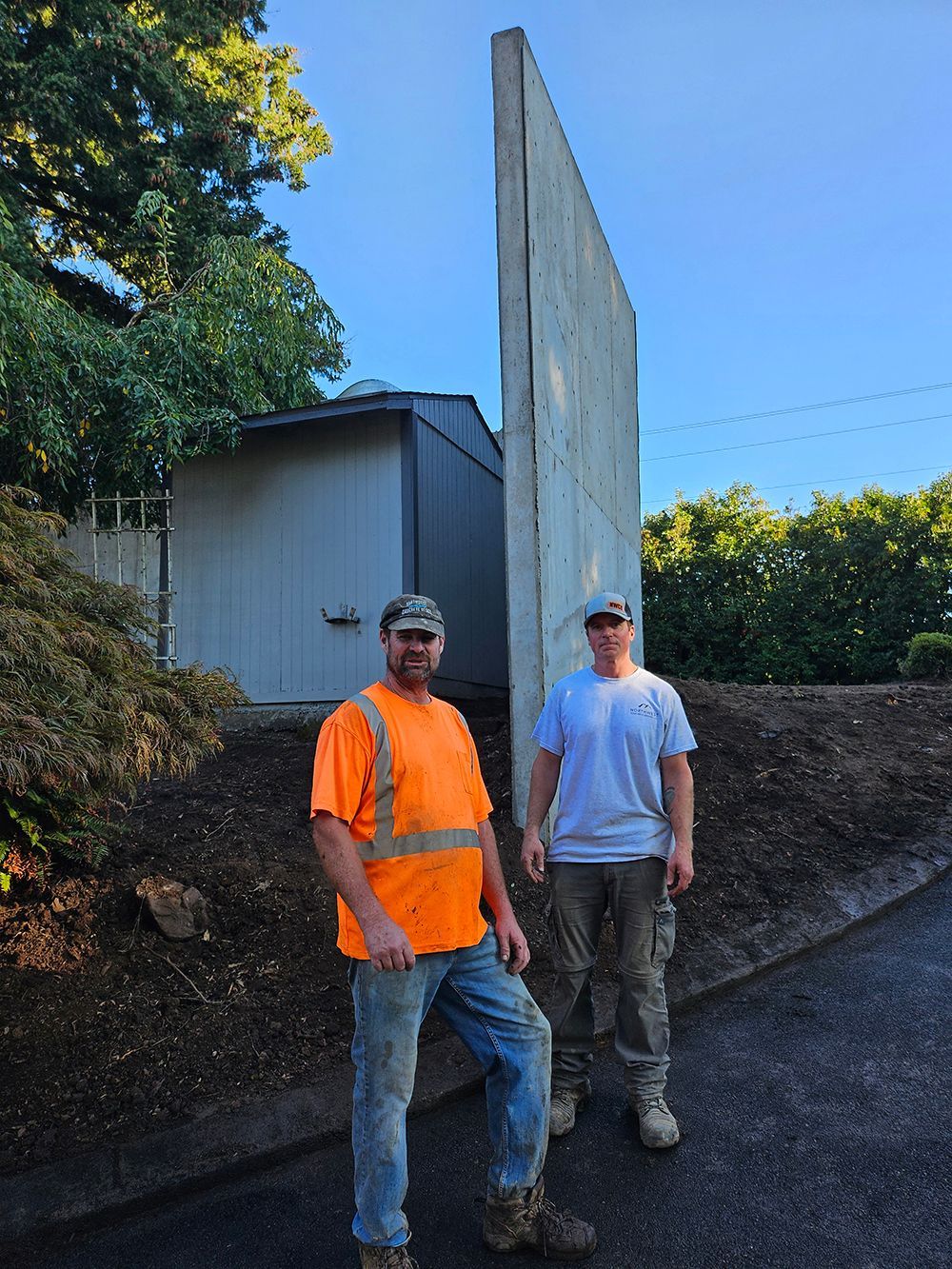 Two men stand by a tall, tilting concrete wall near a shed. One wears an orange vest. Sunny day.