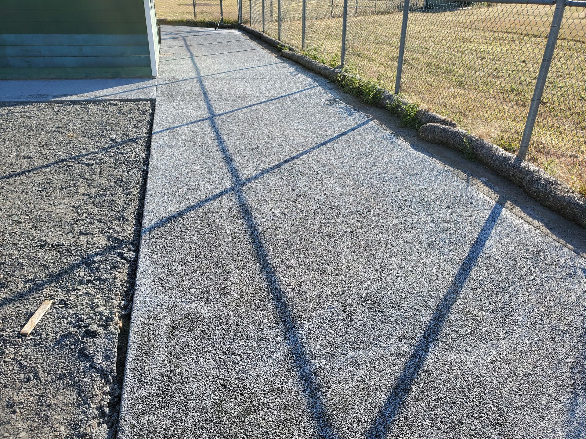 Gravel path next to a fence and a grassy area. The path is in direct sunlight, and shadows are cast on the surface.