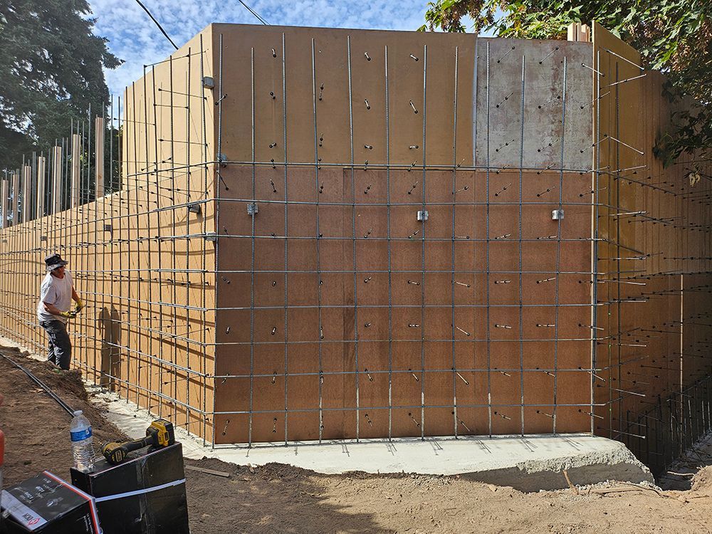 Construction site: Worker by wooden concrete form, with rebar grid. Light brown wood, gray concrete, blue sky.