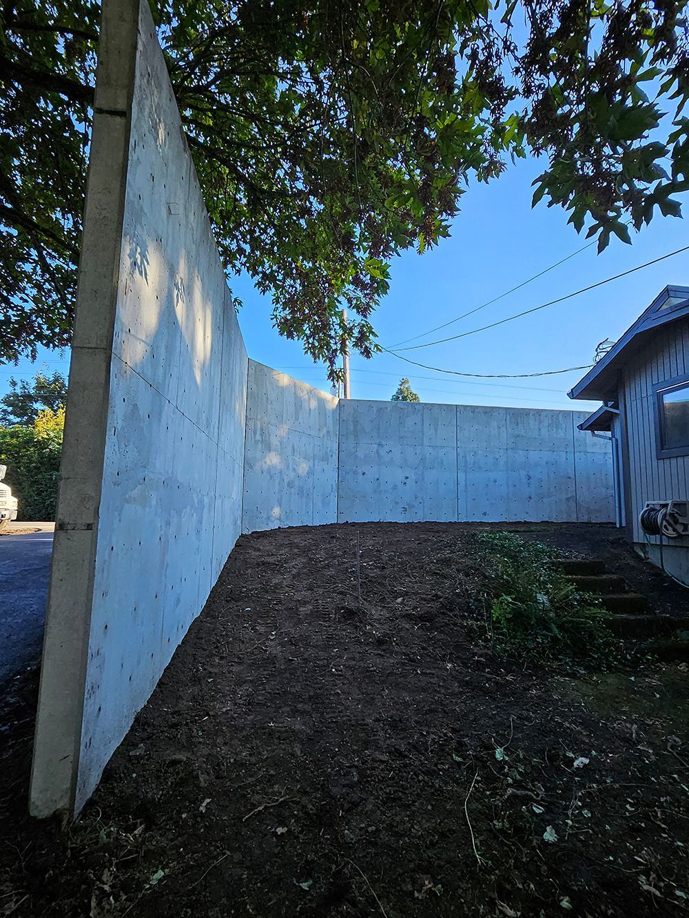 A tall concrete wall curves alongside a house and a tree, with dark soil in the foreground.