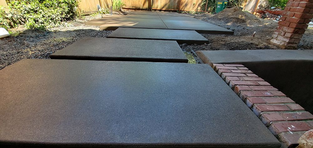 Black stepping stones form a path through a yard with gravel and a red brick wall.