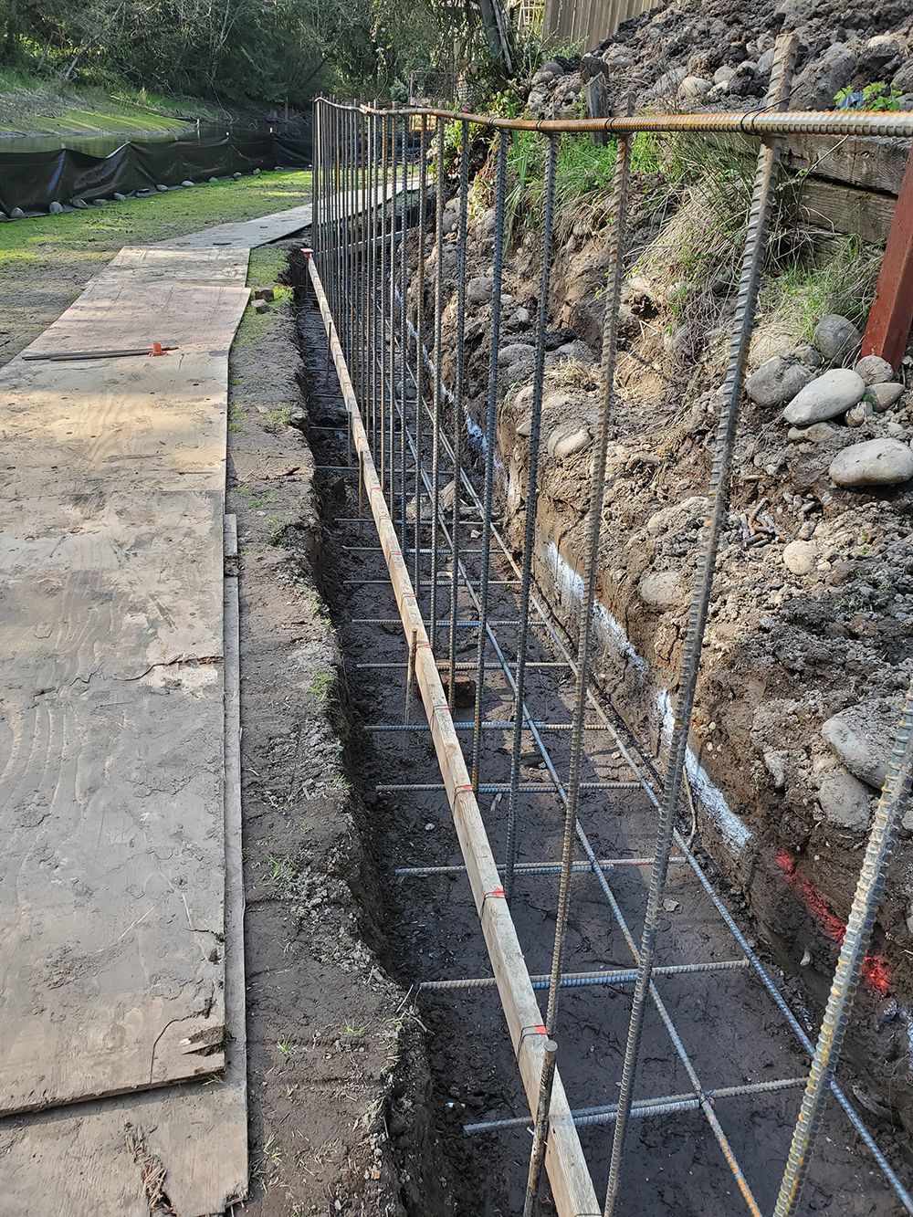 Reinforced steel framework for a retaining wall in a dirt trench, near a pathway and hillside.