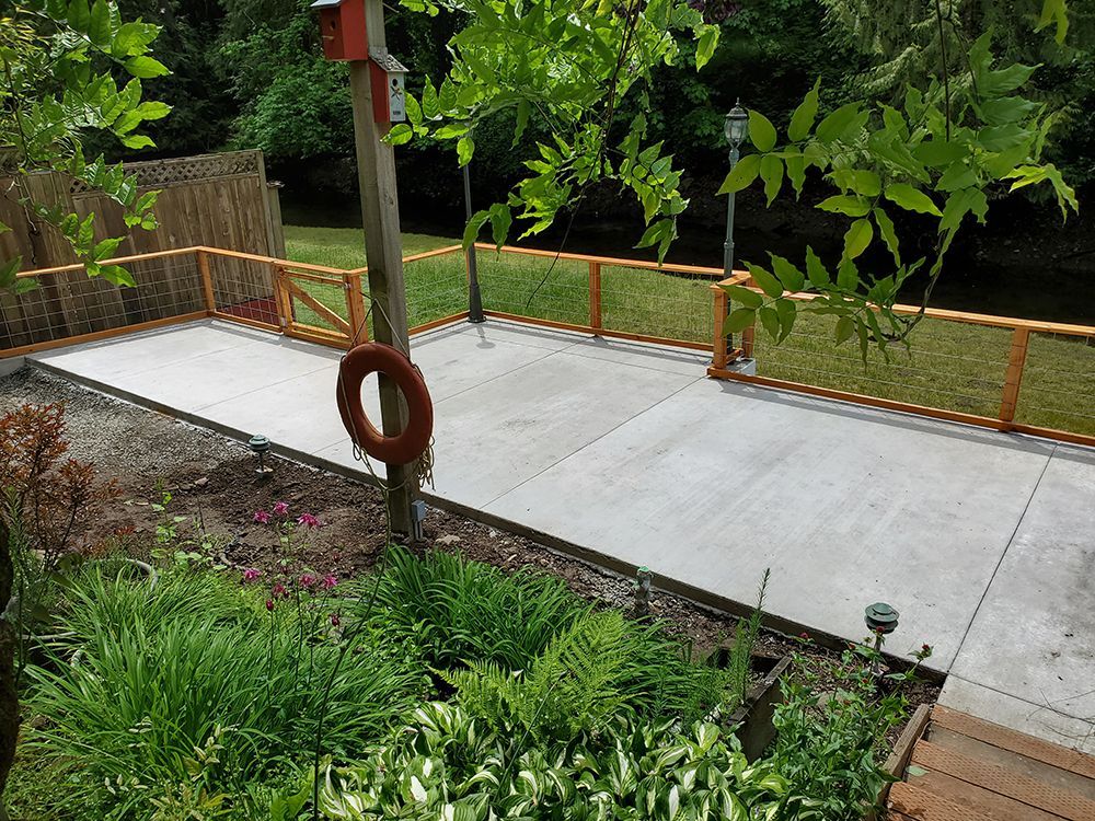 Concrete patio with wooden railing, surrounded by grass and greenery. A life preserver hangs on a pole.