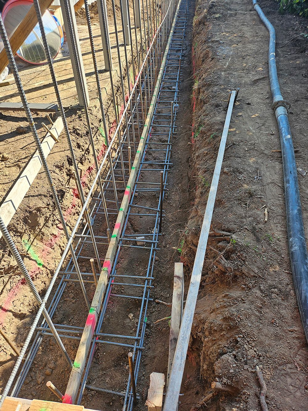 Steel reinforcement bars in a trench, ready for concrete. Wooden forms and a hose are nearby.