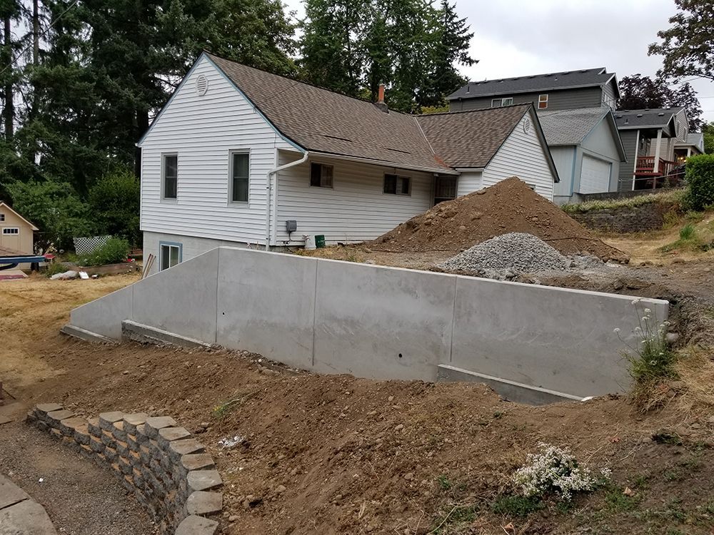 Concrete retaining wall in front of a white house on a sloped lot under construction. Brown dirt, rock pile visible.
