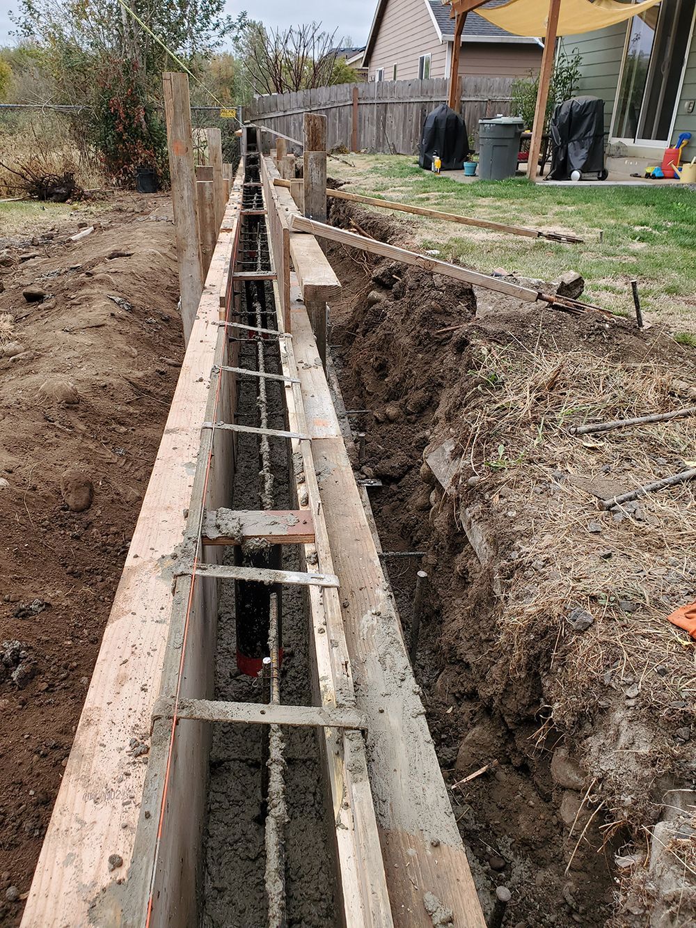 Wooden forms with rebar in a trench, likely for a concrete foundation, in a backyard setting.