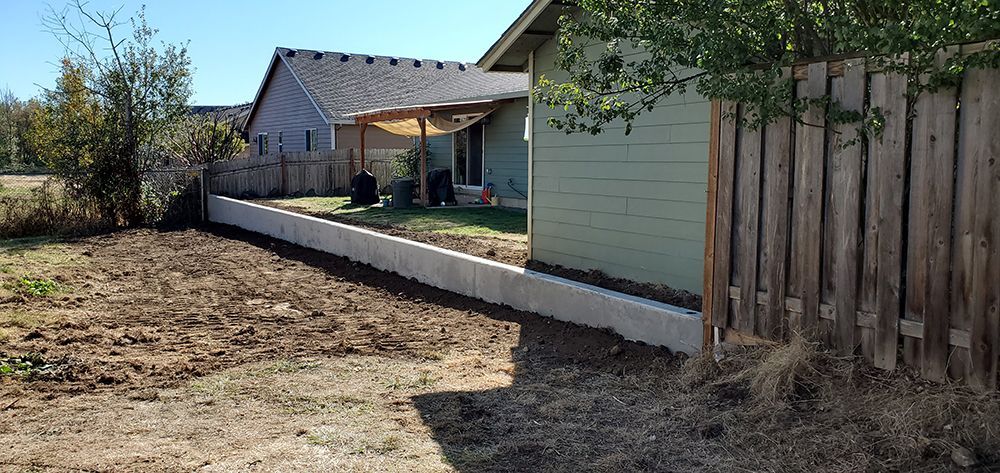 Backyard with a retaining wall, house, and wooden fence. The ground is dirt and a small tree is visible.