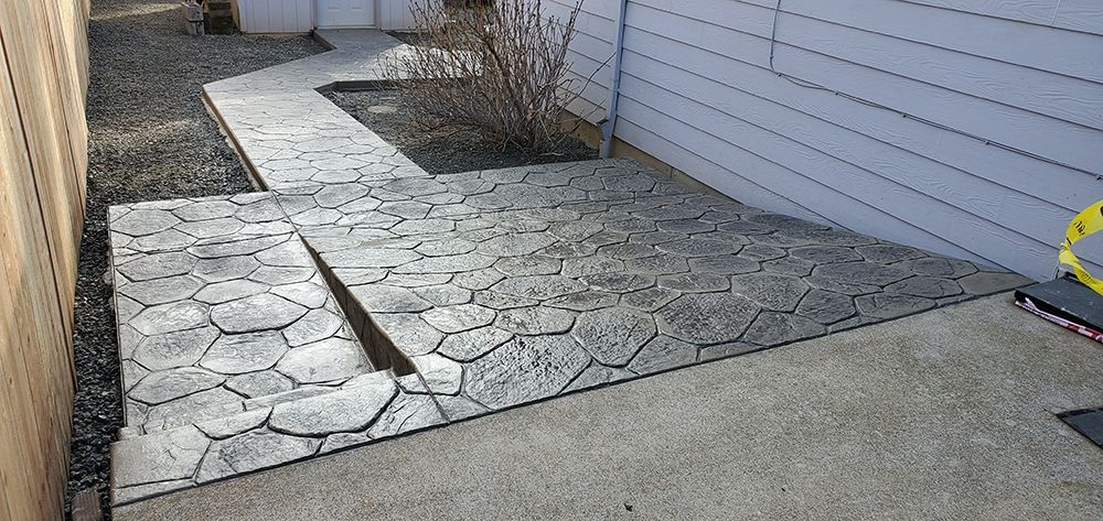 Concrete walkway with textured stones, leading to a plain concrete area next to a blue house and fence.